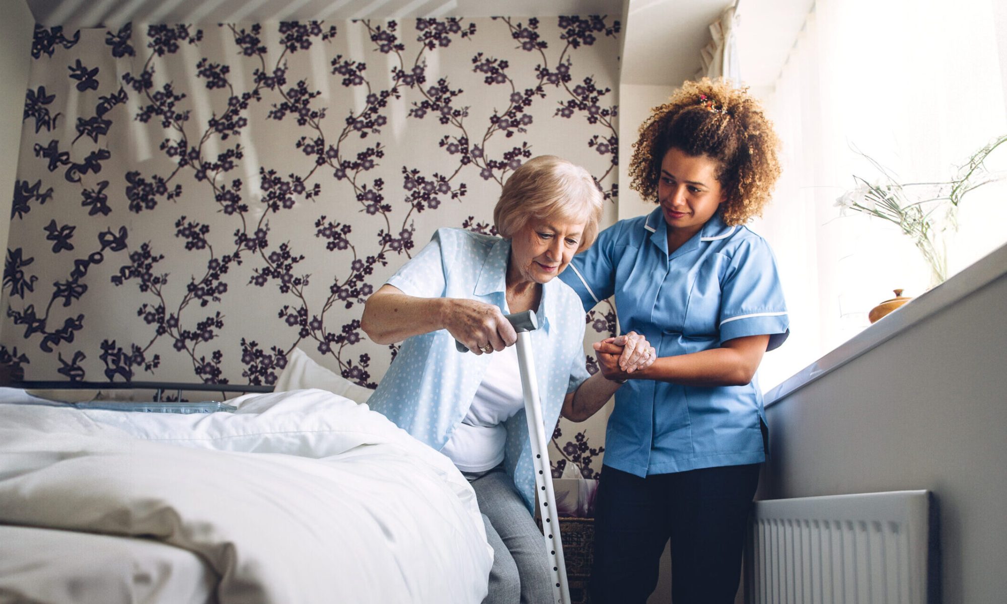 Home Caregiver helping a senior woman get up from her bed with a walking stick.